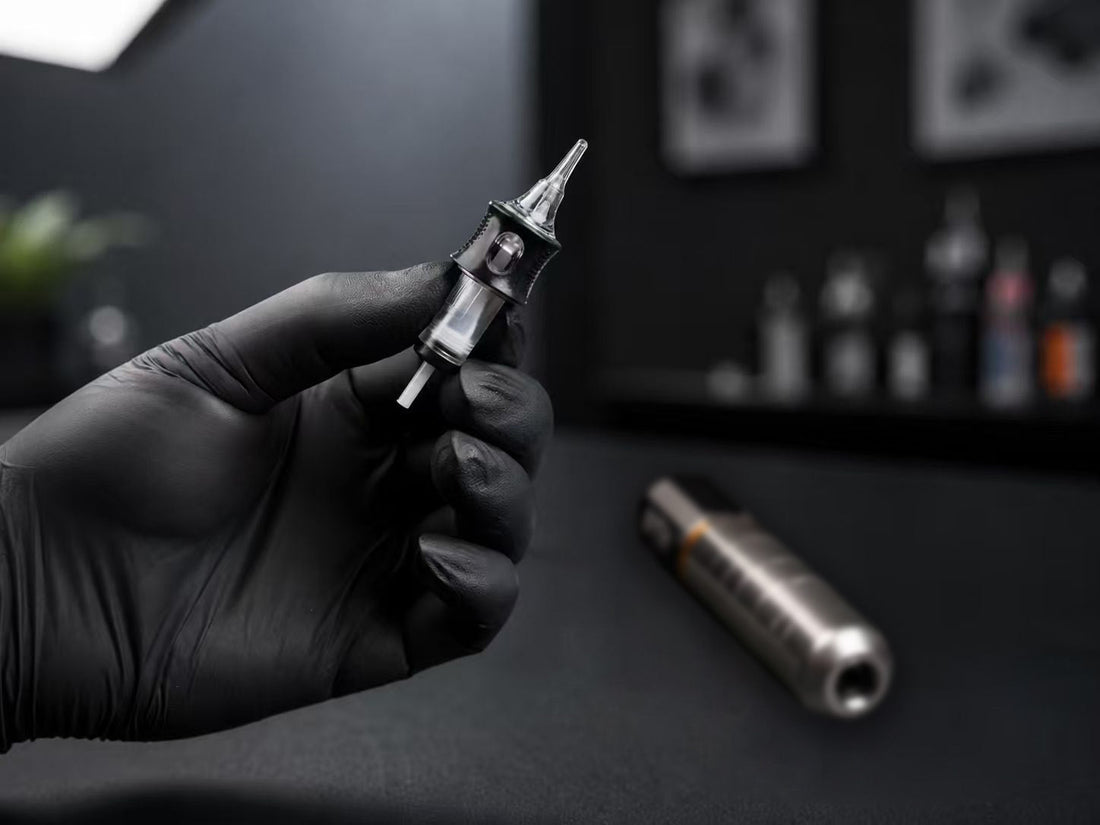 Dragoart Frottage tattoo cartridge needle held by a gloved artist with a tattoo machine in the background.