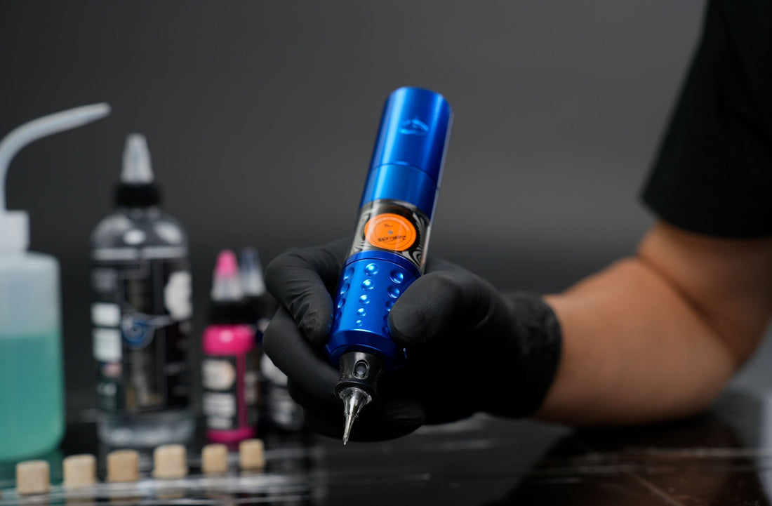 Tattoo artist wearing black gloves holding a blue Dragoart T2 rotary tattoo machine in a professional studio setup with tattoo ink and supplies in the background.