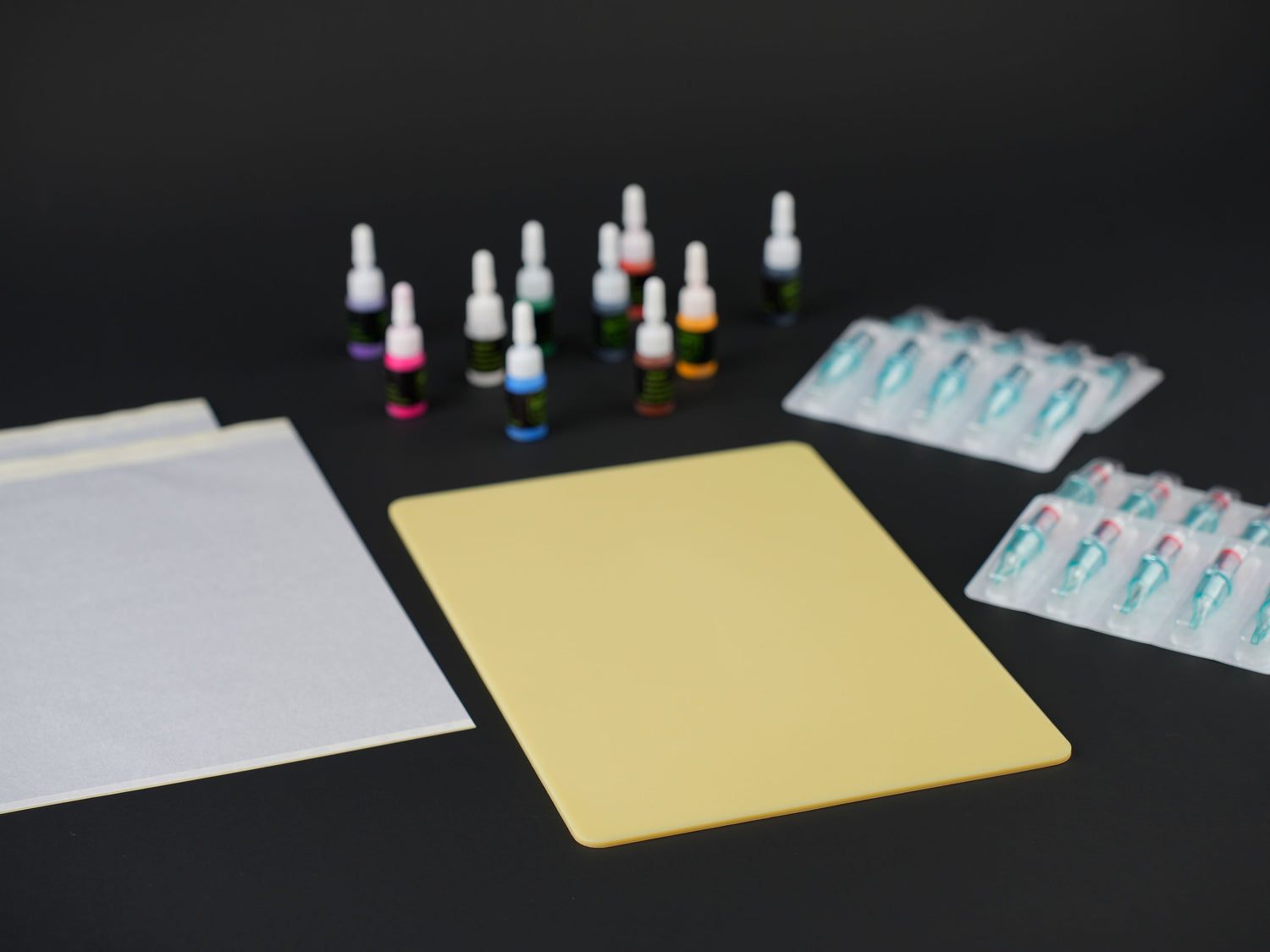 Tattoo kit supplies including practice skin sheet, stencil transfer papers, colorful tattoo inks, and sterile cartridge needles on black background.