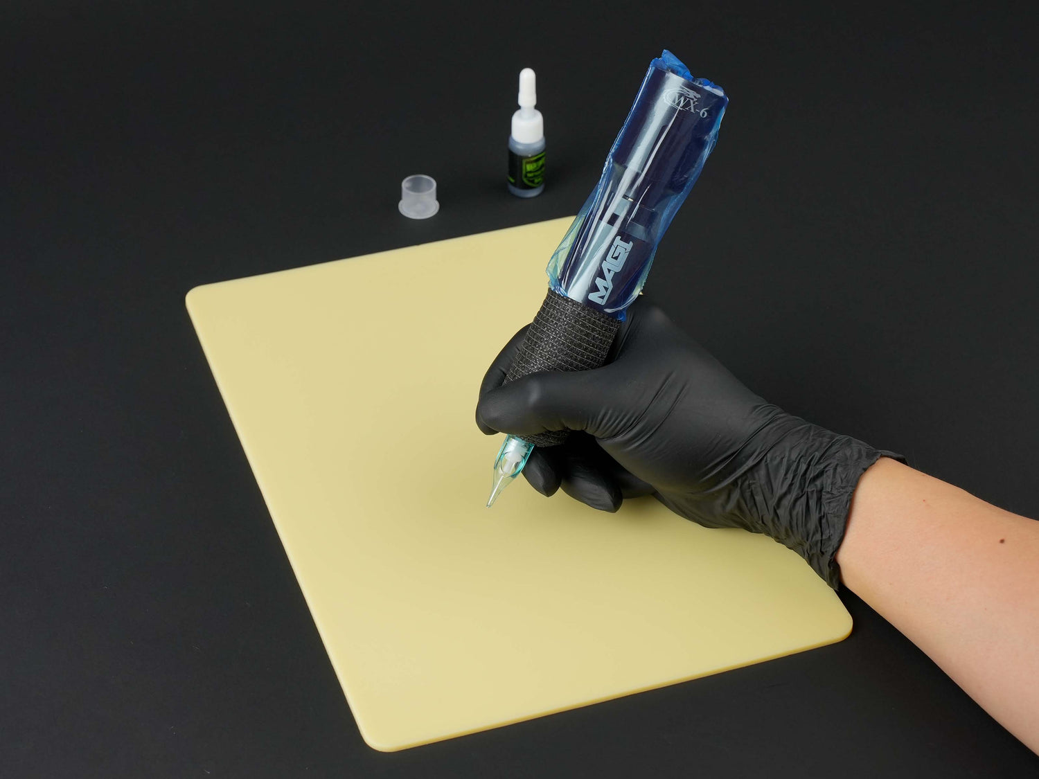Tattoo artist hand in black glove holding MAGI rotary tattoo pen with WX-6 wireless battery, practicing on yellow tattoo practice skin sheet.