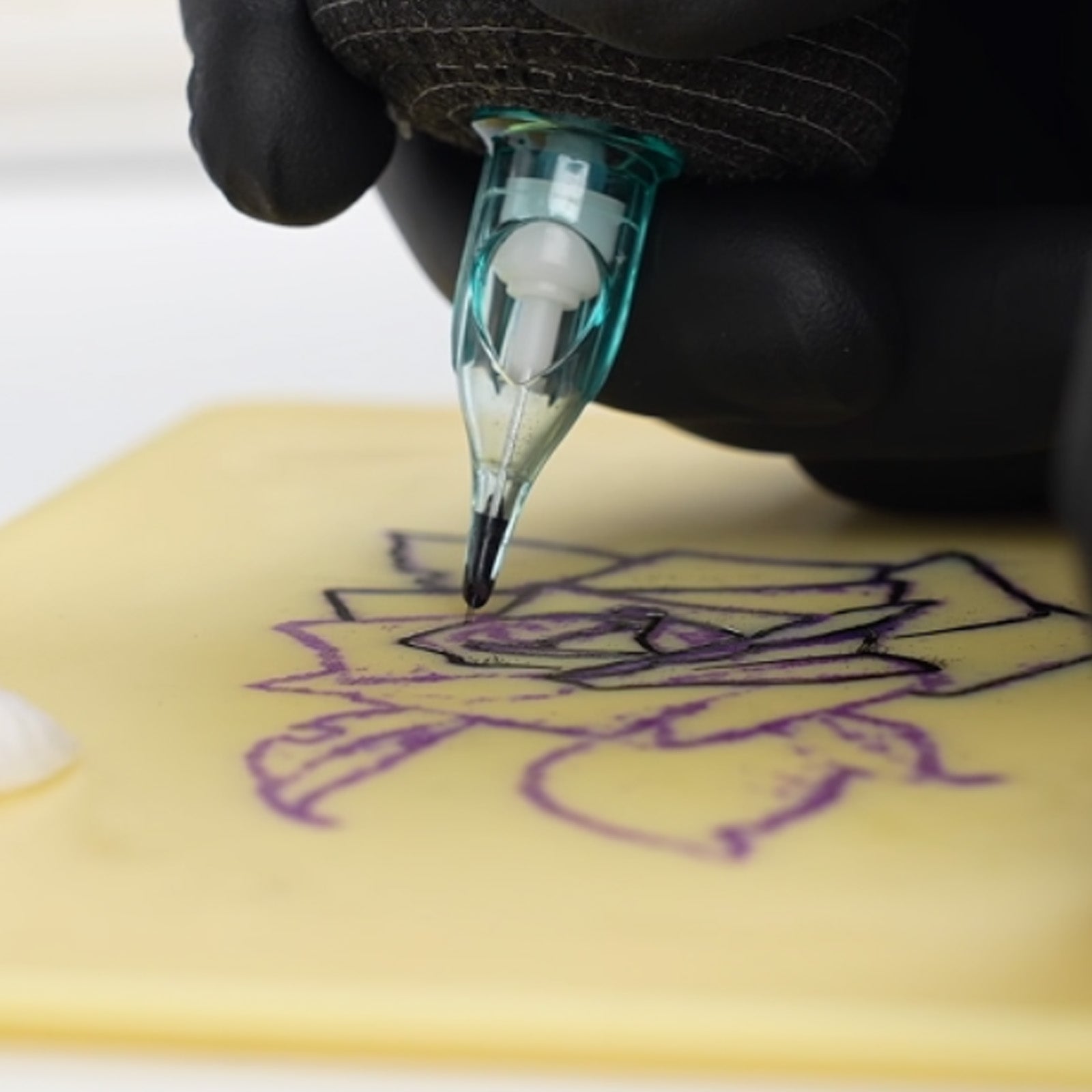 Tattoo artist using cartridge needle on fake practice skin to outline floral tattoo design over stencil