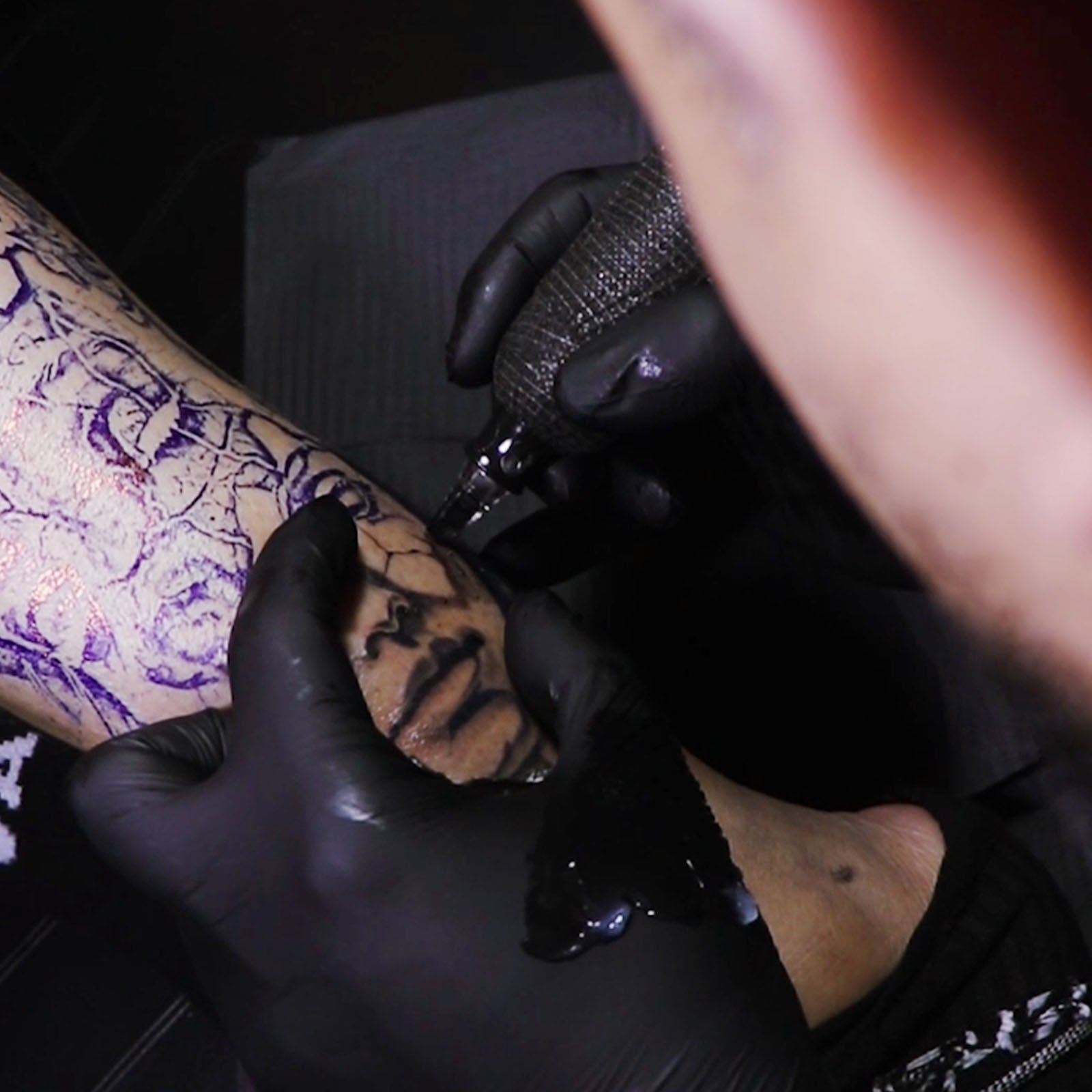 Tattoo artist wearing black nitrite gloves tattooing a detailed black ink design on a client's arm using a professional tattoo machine